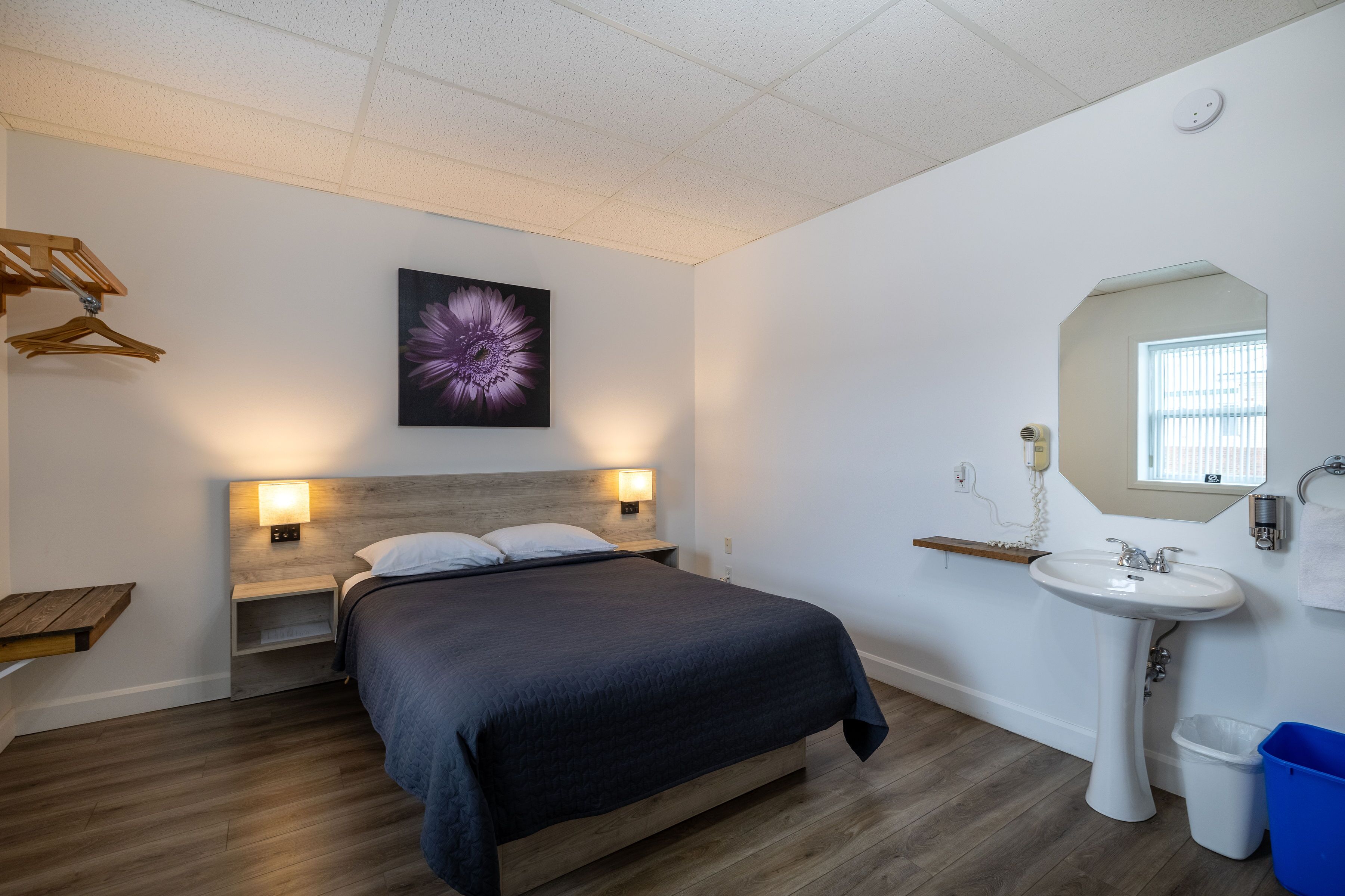 A bedroom with a bed, bedside lamps, a wooden headboard, a wall-mounted artwork, a window, and a bathroom with a sink and mirror.