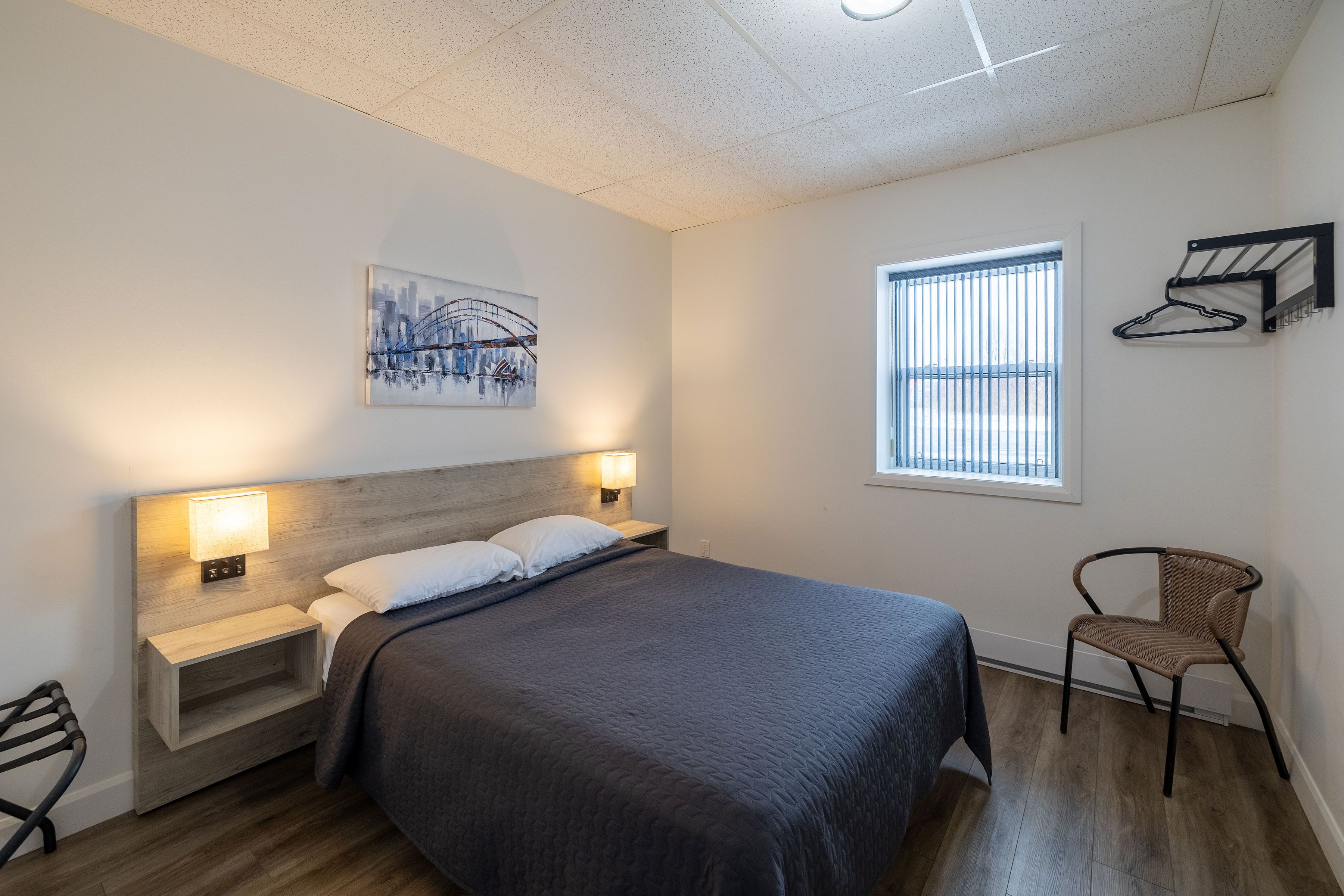 Un dormitorio ordenado con cama, lámparas de noche, una silla, una ventana con persianas y un perchero en la pared.