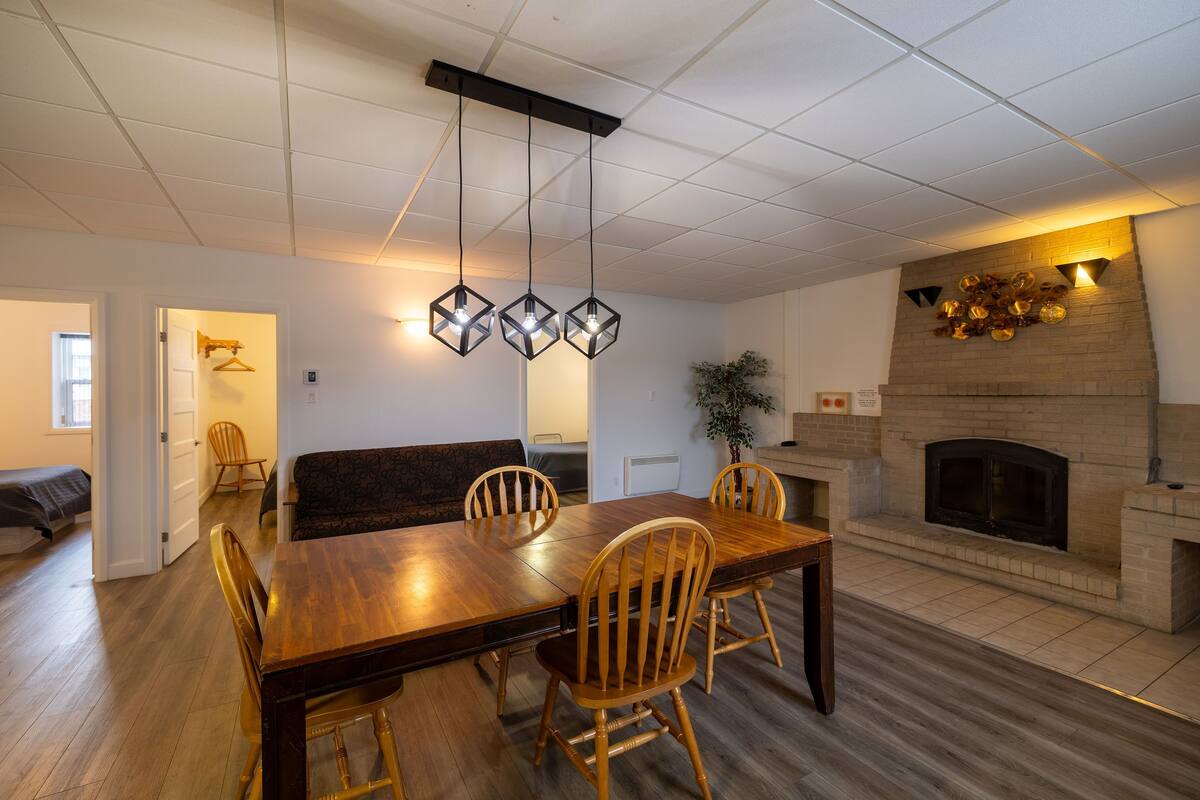 A modern dining area with a wooden table and chairs, a hanging light fixture, and a brick fireplace.