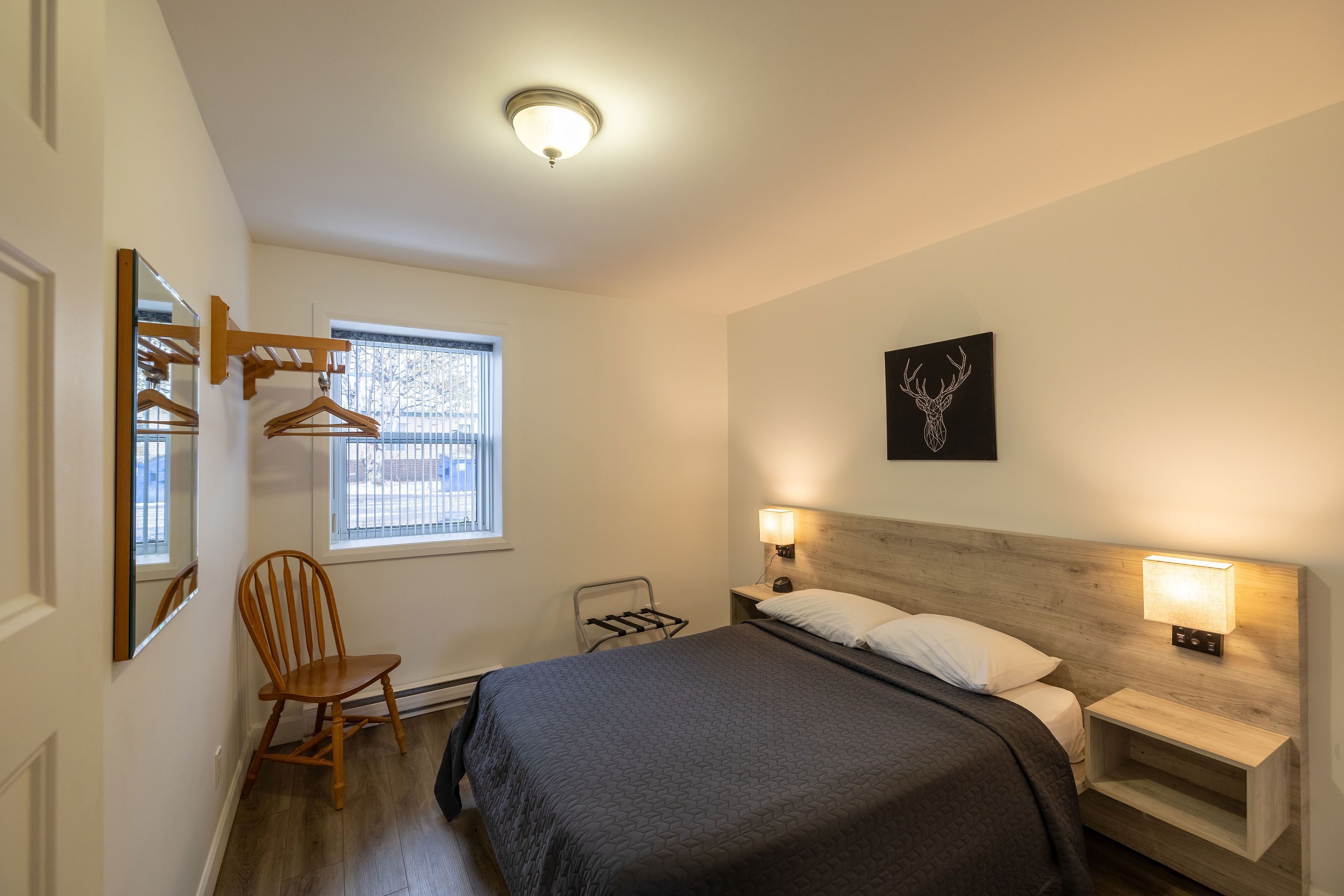 A bedroom with a bed, bedside lamps, a wooden headboard, a wall-mounted shelf, a window with blinds, a wooden chair, and a mirror.
