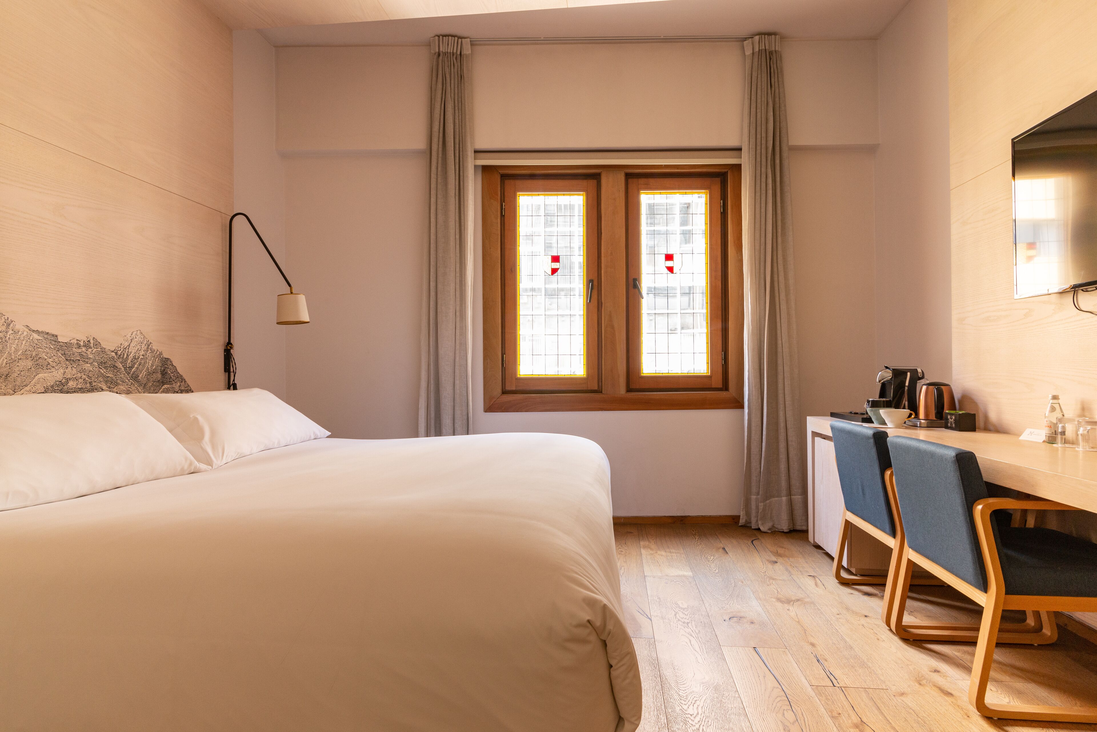 A hotel room with a bed, a desk, two chairs, a lamp, a window with curtains, and a television on the wall.