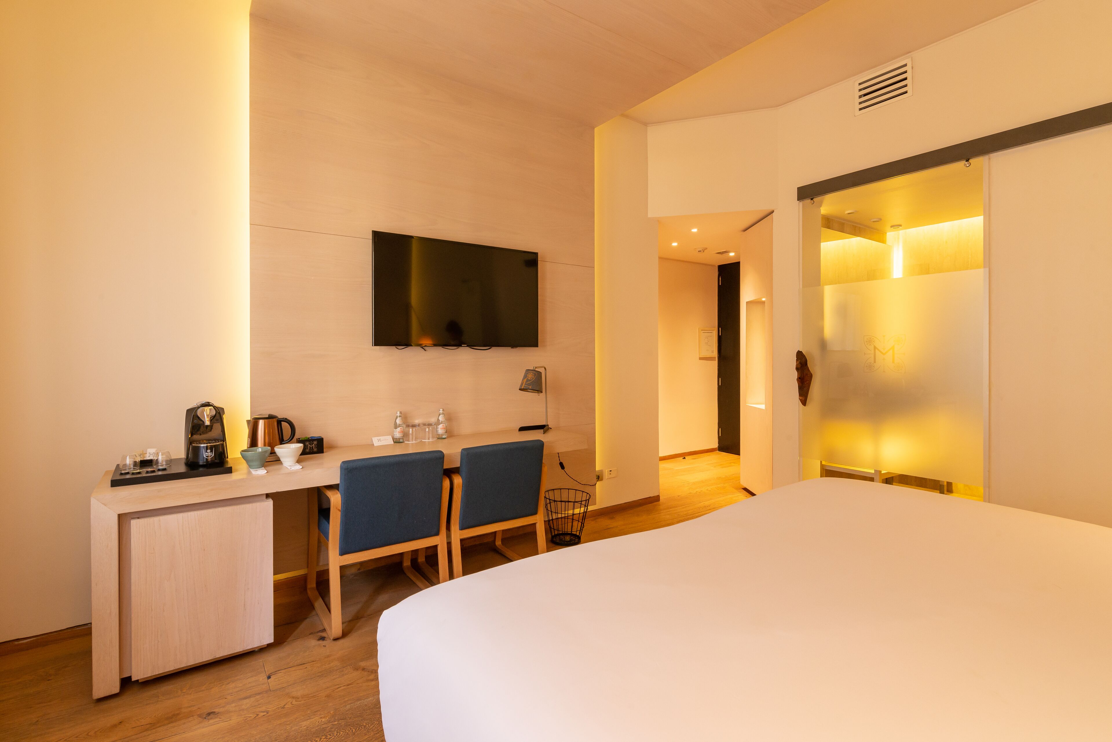 A modern hotel room with a bed, a desk with chairs, a television, and a sliding door.