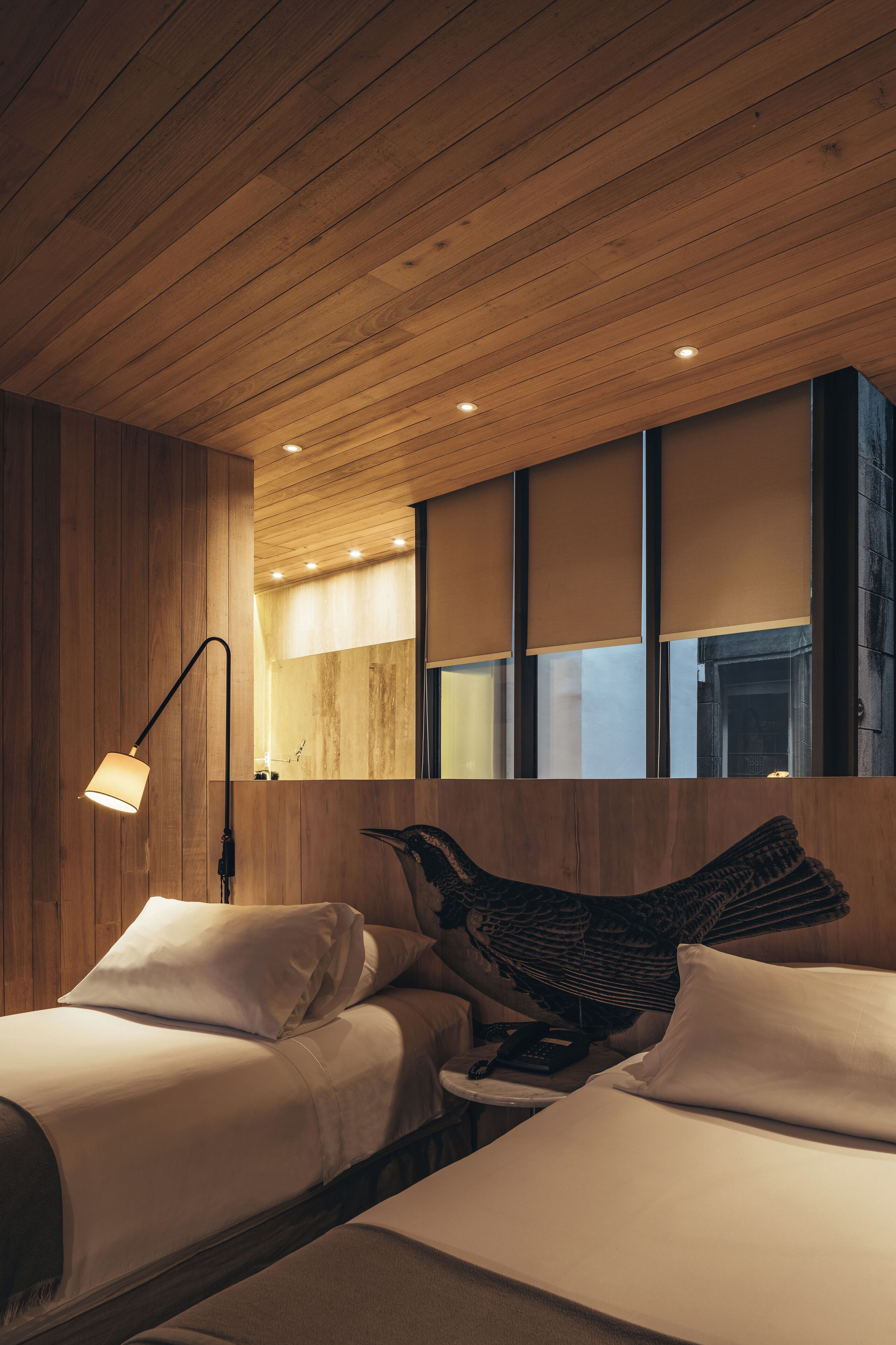 A hotel room with wooden panel walls, a large bed with white bedding, a bedside lamp, and a view of the outside through a window with blinds.