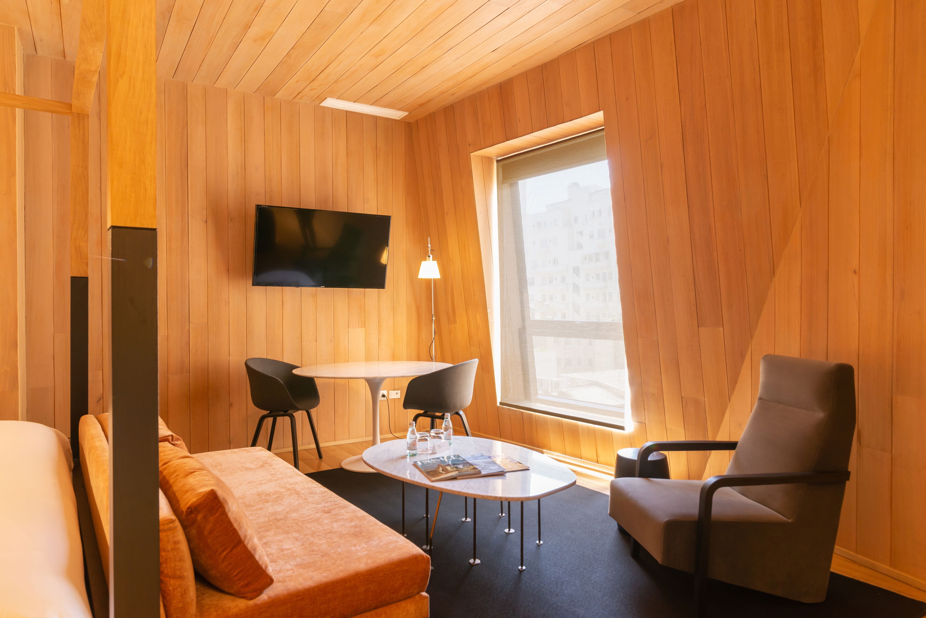 Una habitación moderna con interior de madera, un televisor de pantalla plana montado en la pared, una mesa redonda con sillas y un gran ventanal que proporciona luz natural.