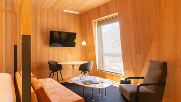 Una habitación moderna con interior de madera, un televisor de pantalla plana montado en la pared, una mesa redonda con sillas y un gran ventanal que proporciona luz natural.