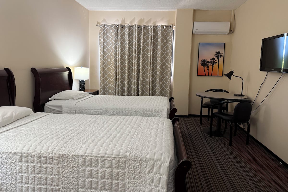 A hotel room with two beds, a desk, a chair, a lamp, an air conditioner, and a painting of palm trees.
