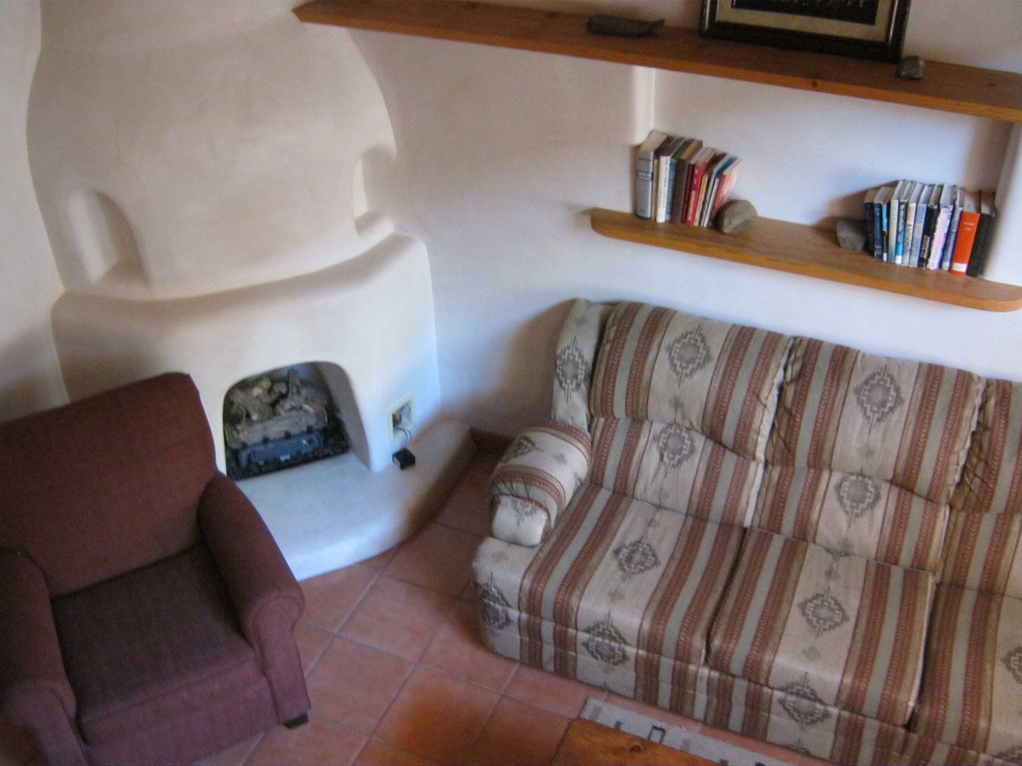 A living room with a red armchair, a patterned sofa, a white cabinet, and a wooden shelf with books.