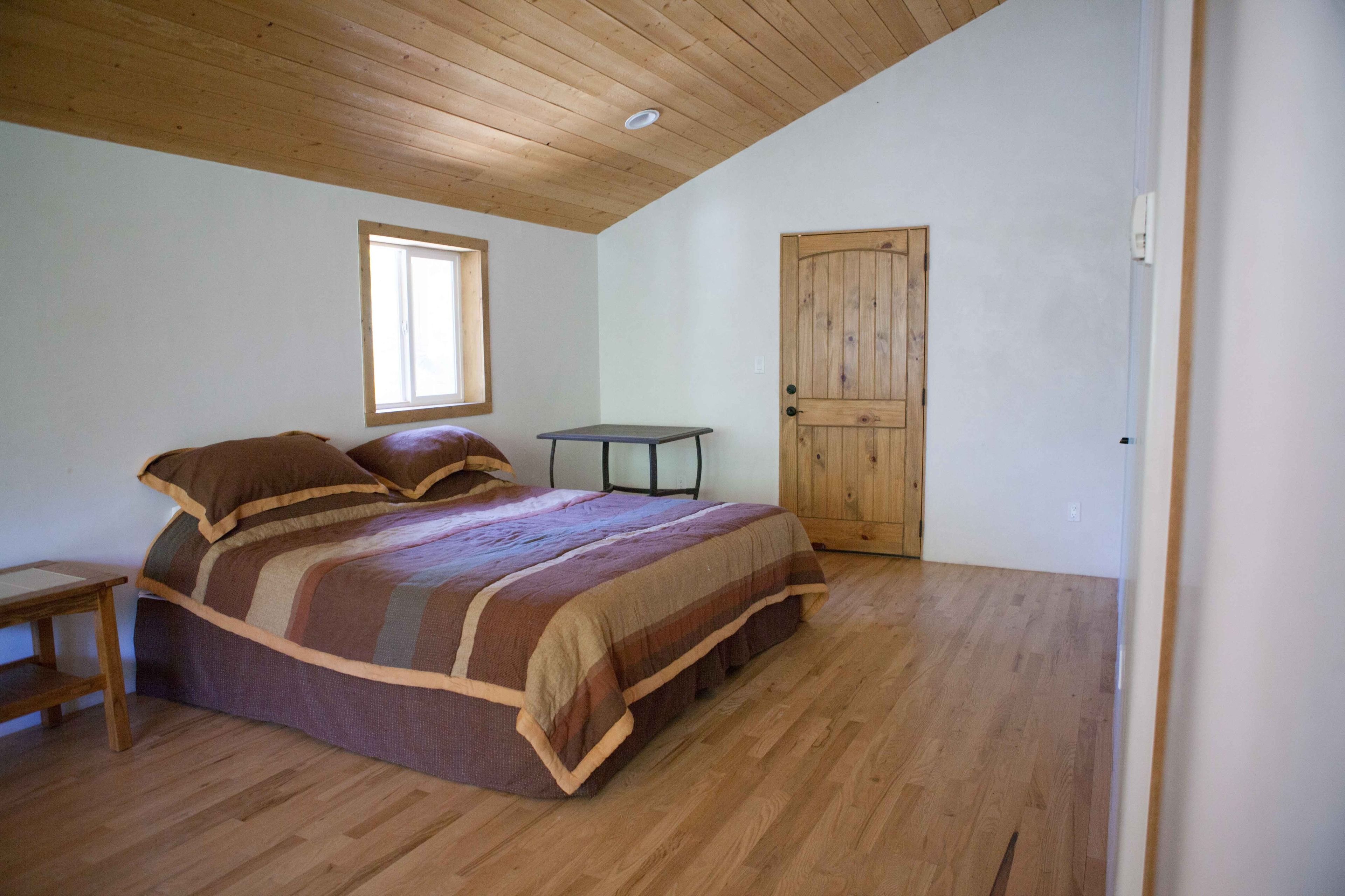 A bedroom with a bed, wooden floor, a window, a door, and a small table.