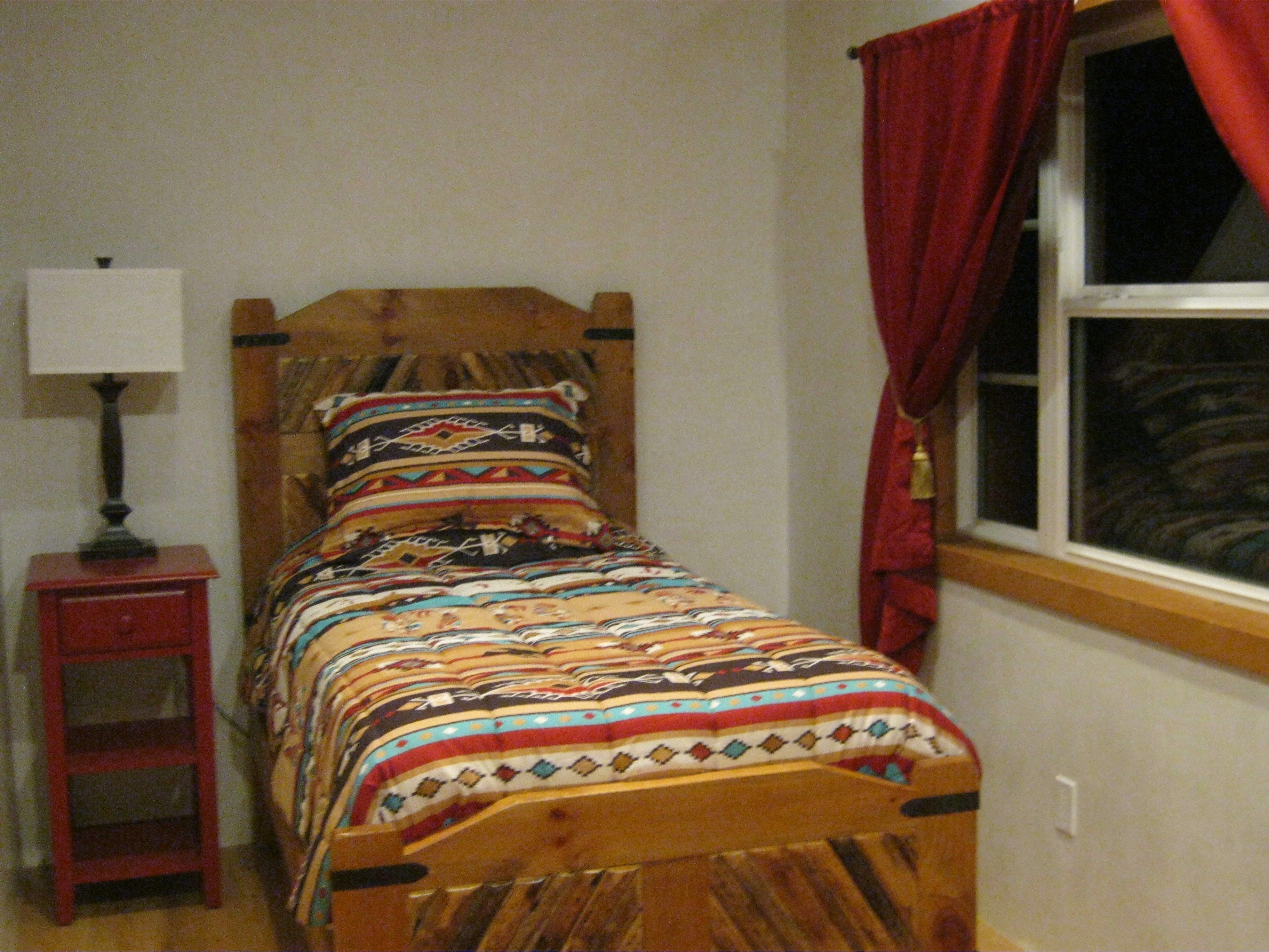 Una cama de madera con colcha estampada, una mesita de noche con lámpara y una ventana con cortinas rojas.