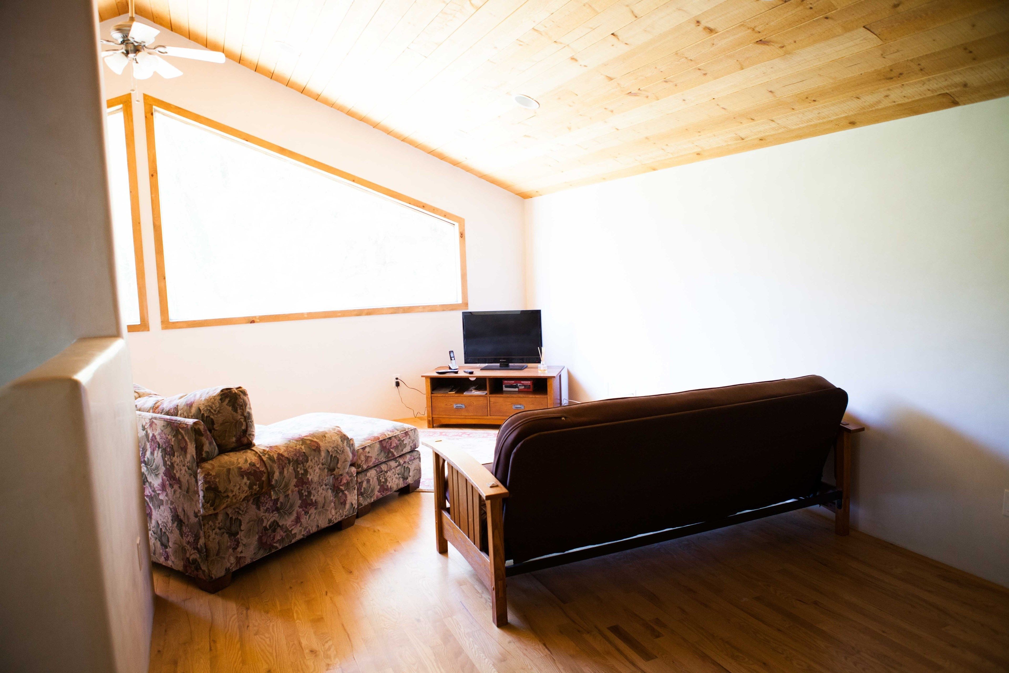 A room with a wooden ceiling, a bed, a sofa, a TV on a wooden cabinet, and a window.