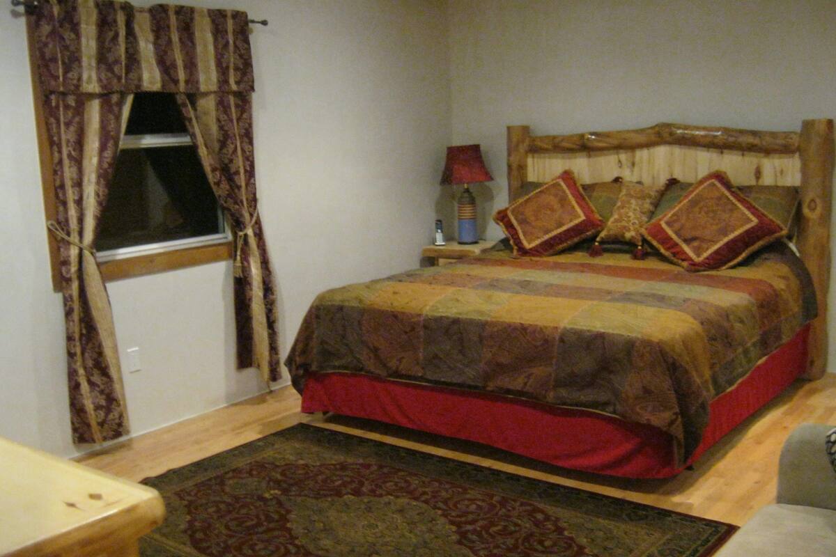 A bedroom with a wooden bed, a nightstand with a lamp, a window with curtains, and a patterned rug.