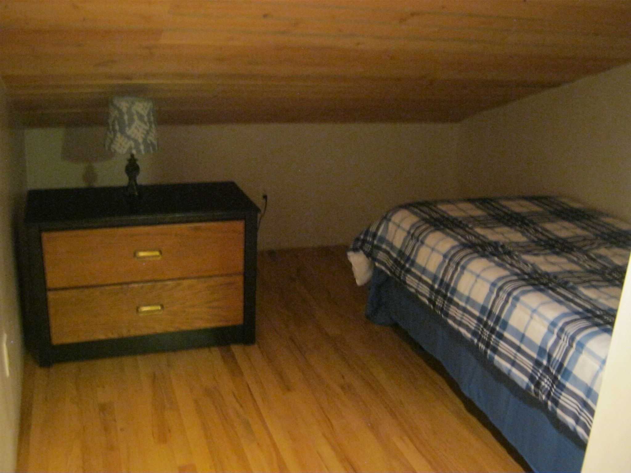 A bedroom with a wooden dresser, a bed with a plaid blanket, and a lamp on the dresser.