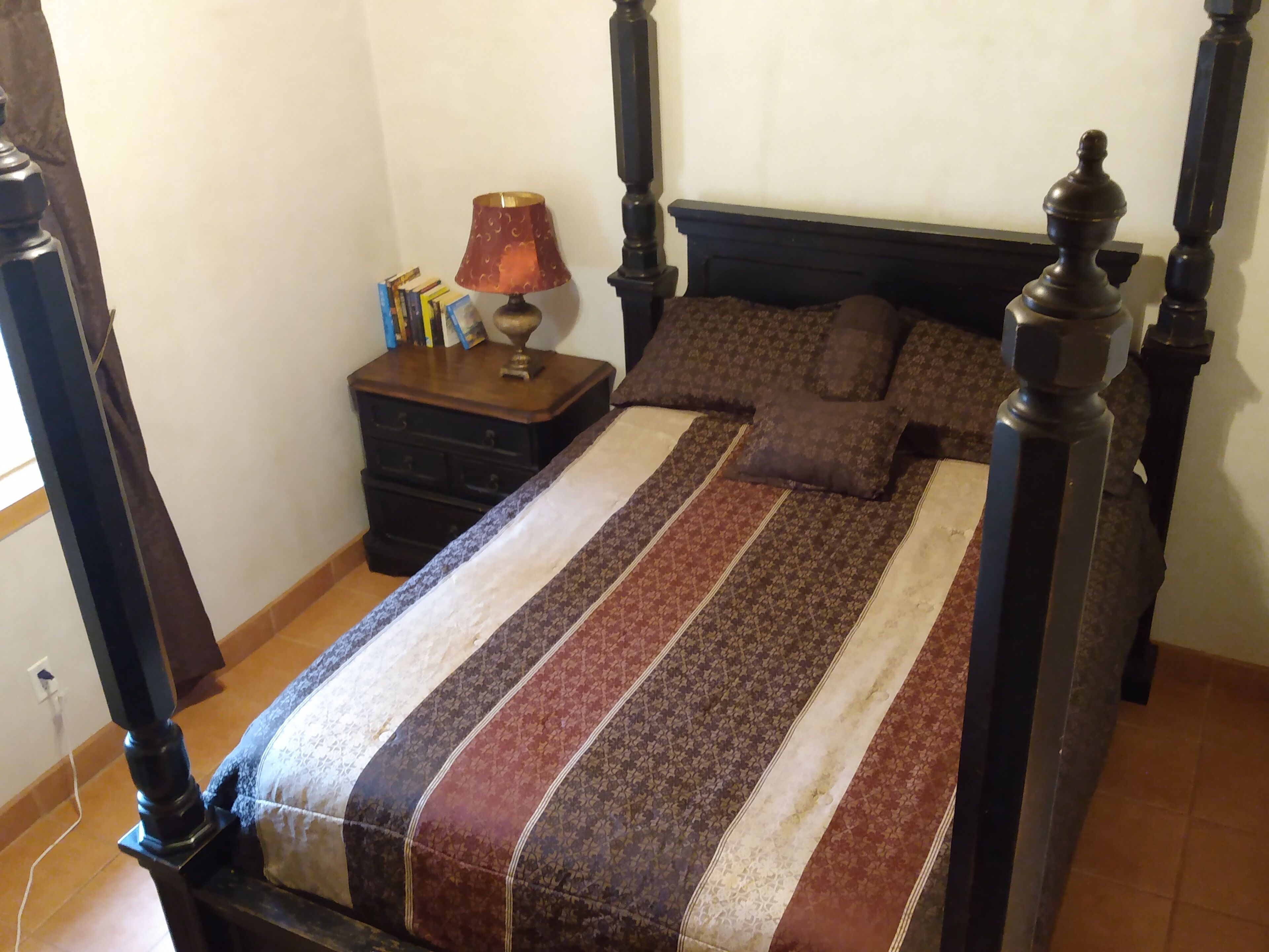 A bedroom with a four-poster bed, a nightstand with books, and a lamp.
