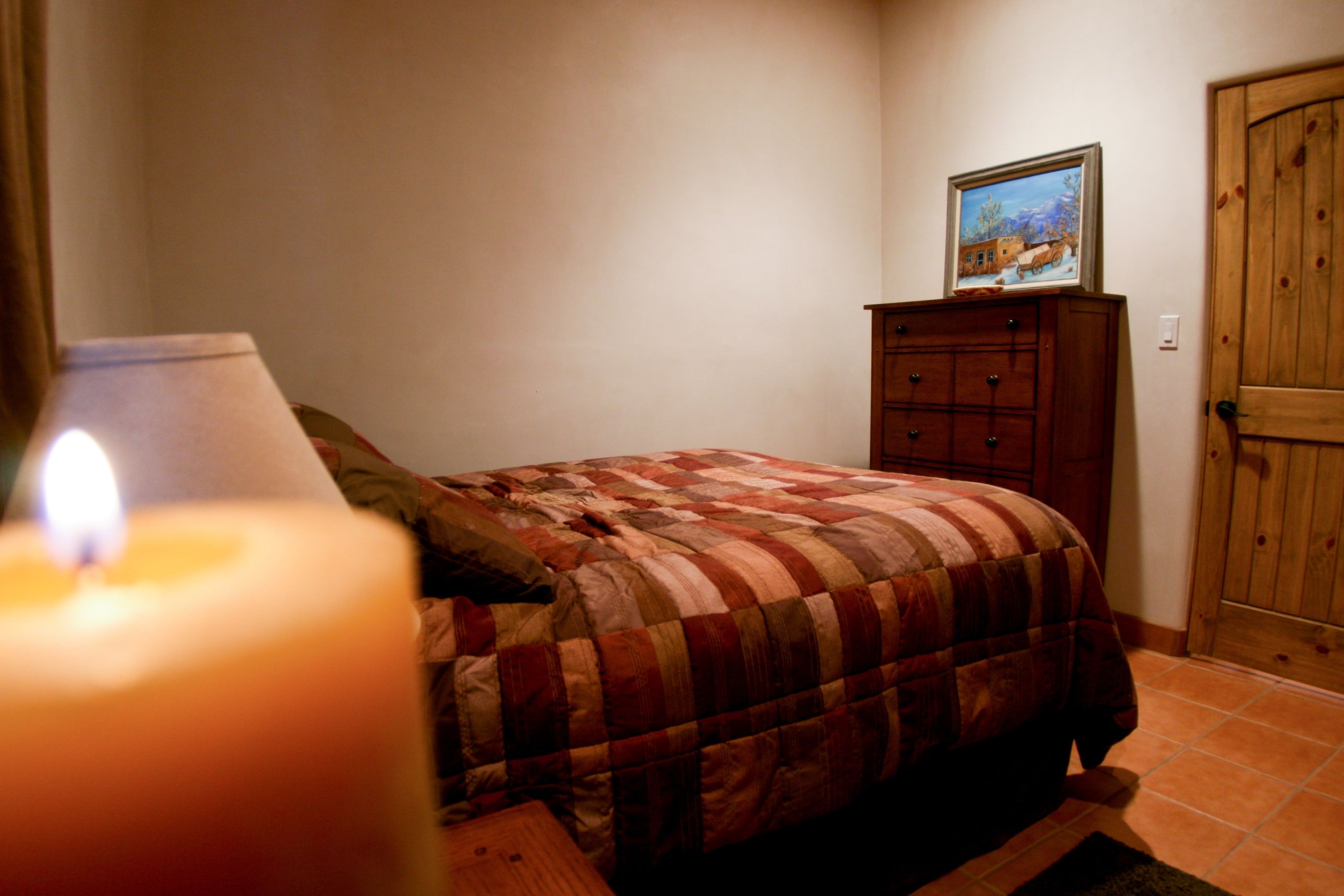 A bedroom with a bed, a wooden dresser, a painting on the wall, and a lit candle.
