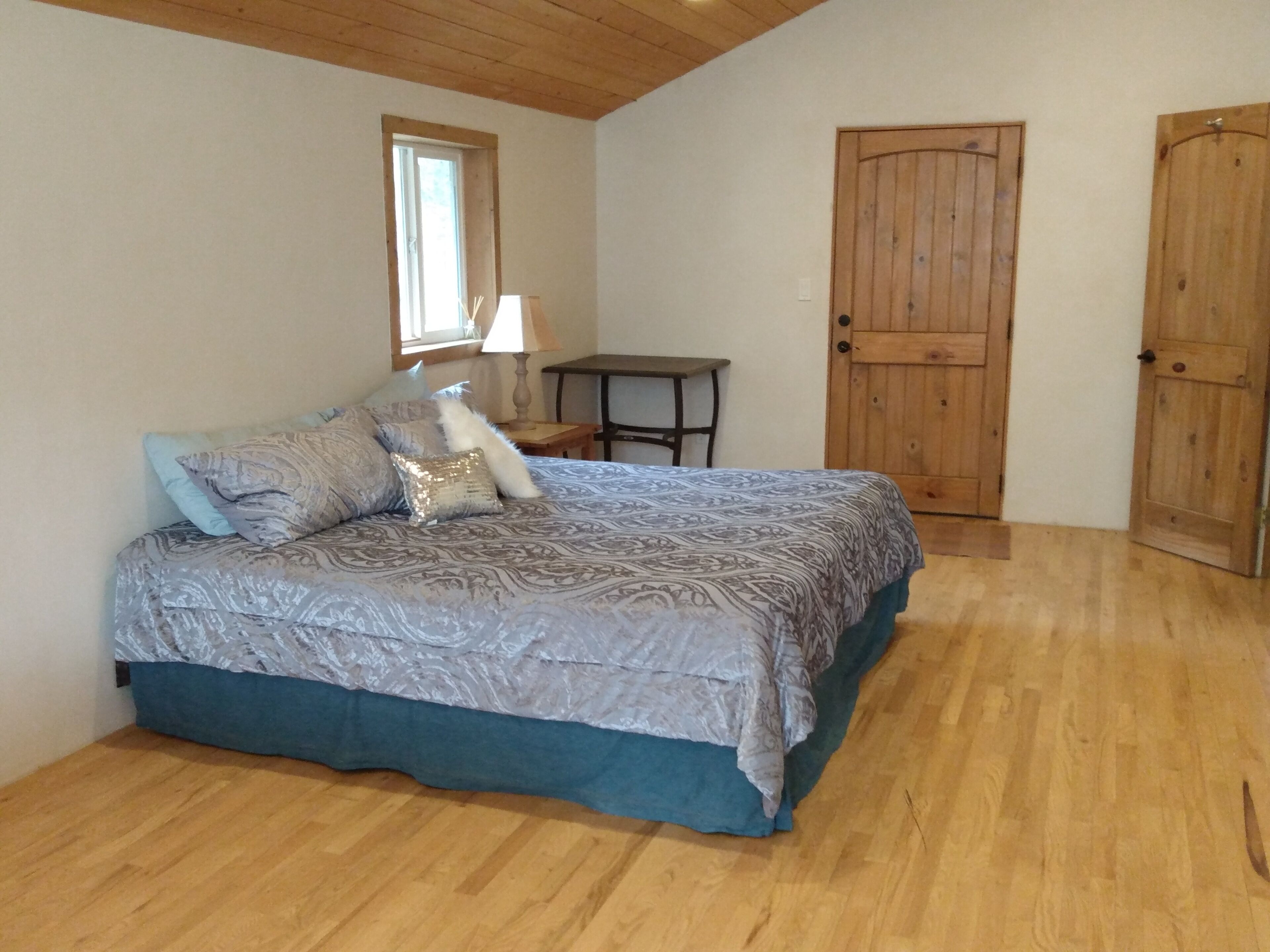 Un dormitorio con cama, piso de madera, una ventana, una mesa y dos puertas de madera.