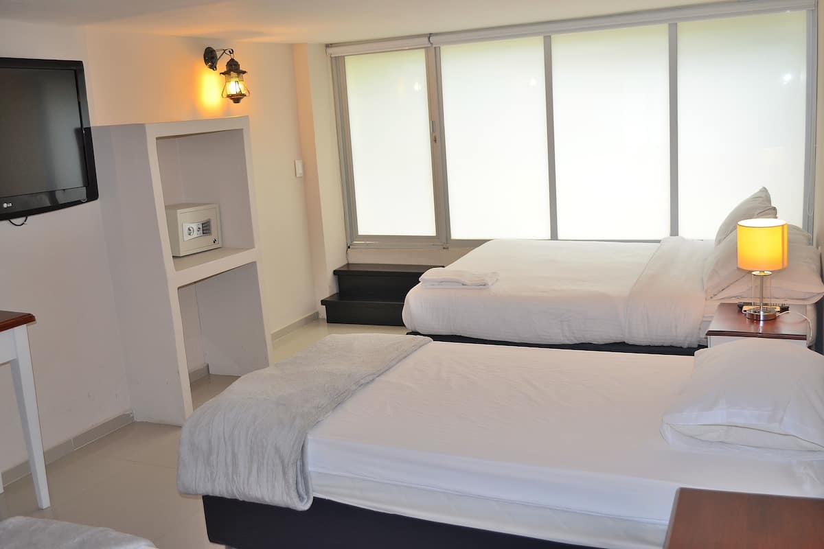 A hotel room with a bed, a TV mounted on the wall, a lamp on the bedside table, and a window with blinds.
