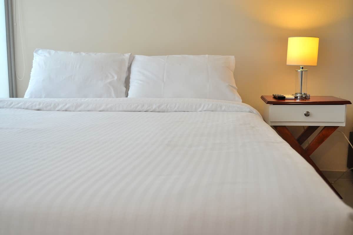 A neatly made bed with white linens, a bedside table with a lamp, and a nightstand.