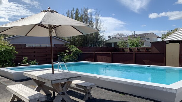 A rectangular swimming pool with a wooden deck and picnic table, surrounded by a wooden fence and greenery. - Colonial Motel (Papakura)