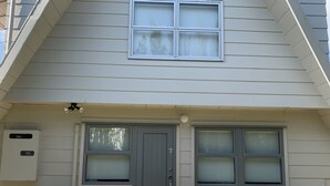 A house with a grey door, two windows with frosted glass, an air conditioning unit, and two white chairs outside. - Colonial Motel (Papakura)