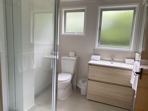 A bathroom with a glass shower enclosure, a toilet, a sink with a mirror, and a window above the sink. - Colonial Motel (Papakura)