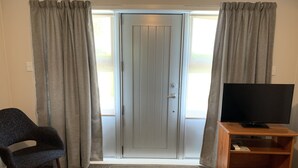 A room with a grey door, grey curtains, a dark blue chair, a flat-screen TV on a wooden stand, and a small wooden shelf. - Colonial Motel (Papakura)