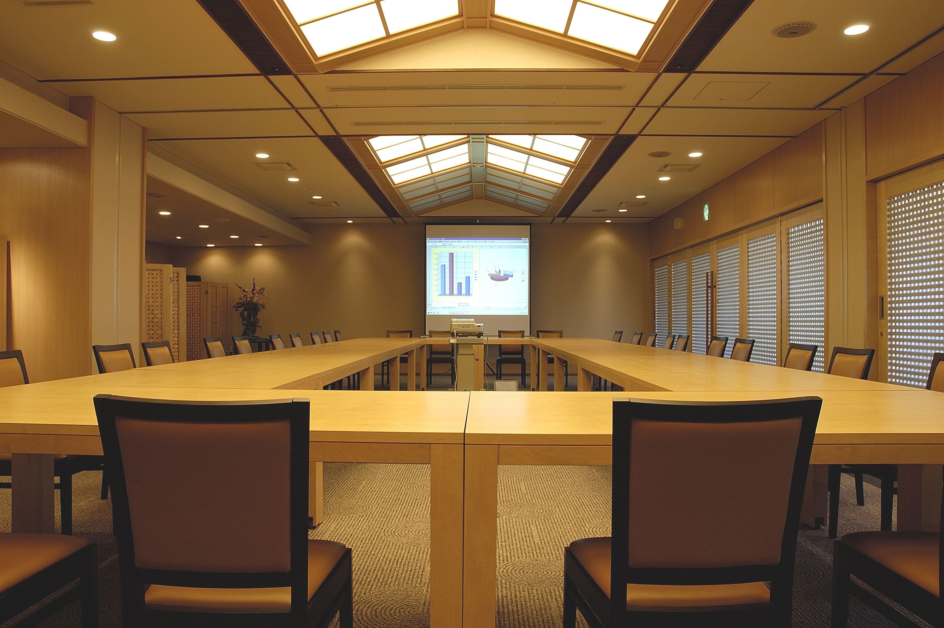 A conference room with a long table, chairs, a projector screen, and a skylight ceiling.