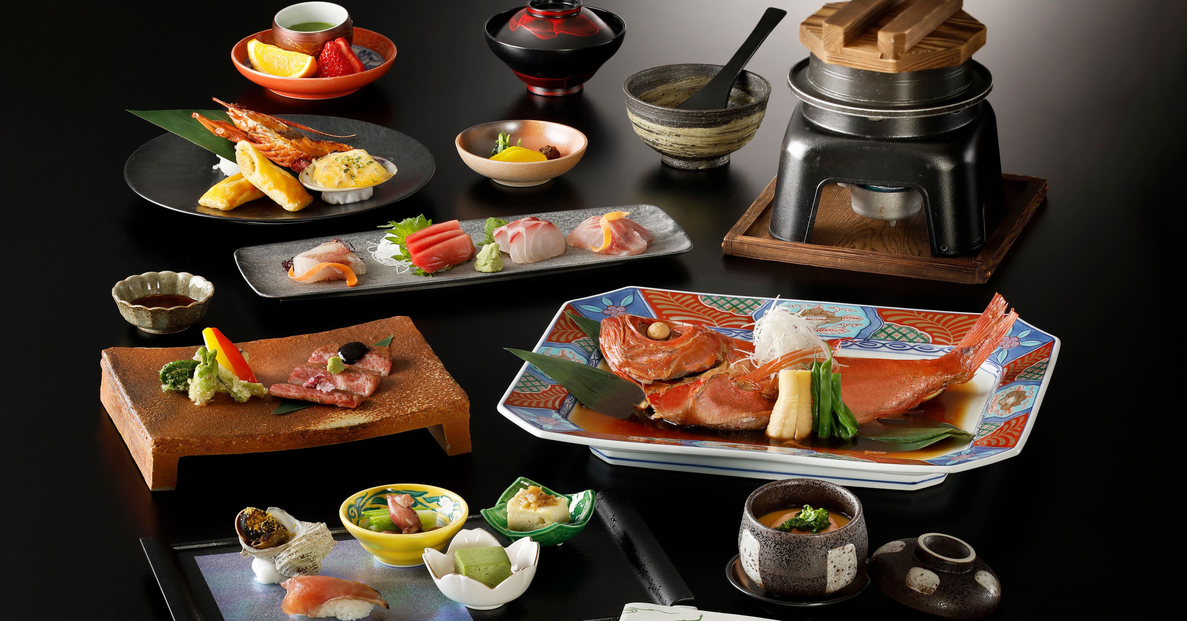 Variedad de platos japoneses, como sushi, sashimi y un cangrejo grande, servidos sobre una mesa negra con cuencos tradicionales y un recipiente para sopas calientes.