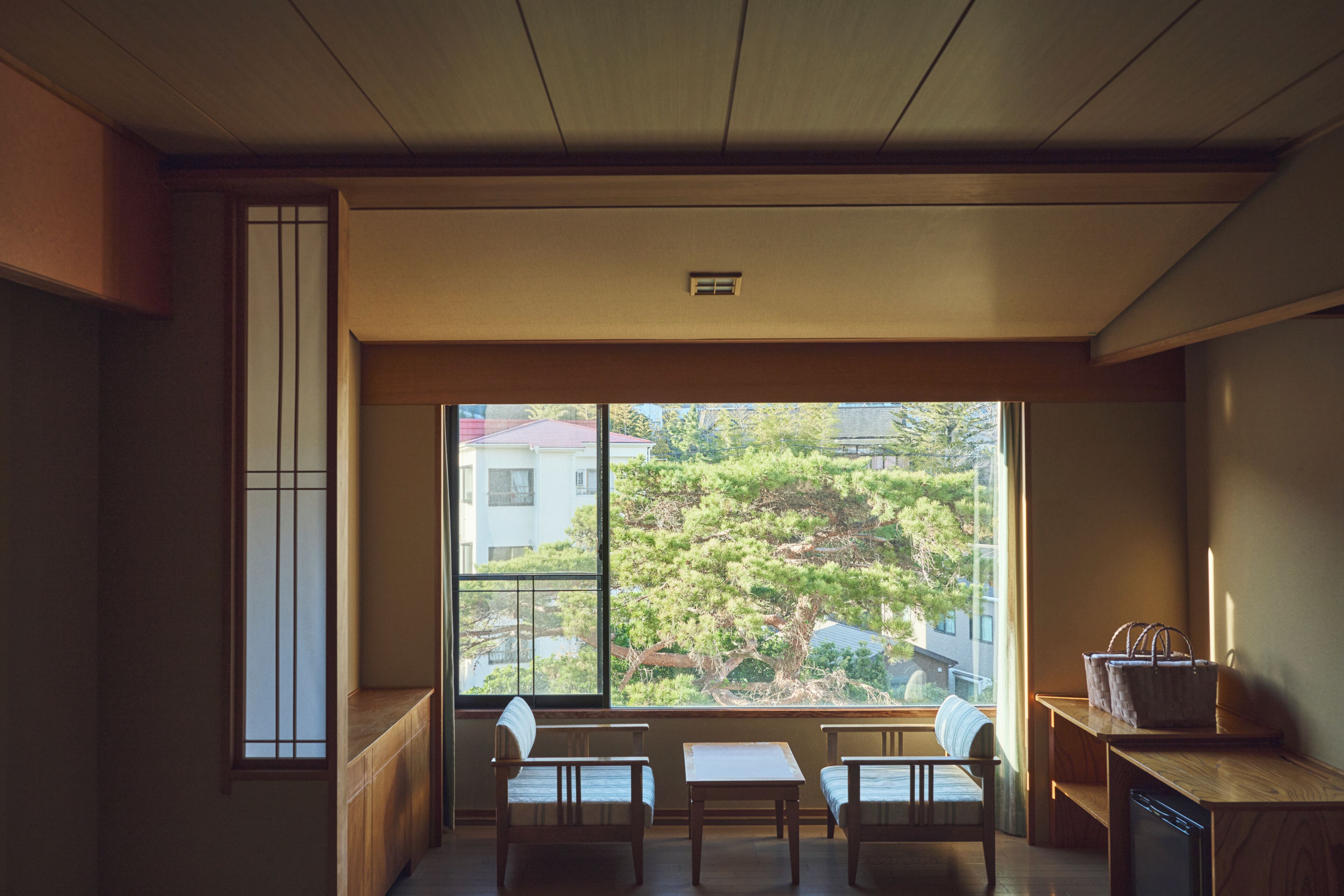 A traditional Japanese room with tatami flooring, a low table, two chairs, and a large window offering a view of trees and buildings.