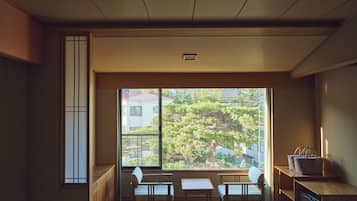 Um quarto japonês tradicional com tatames, uma mesa baixa, duas cadeiras e uma janela grande com vista para árvores e edifícios.