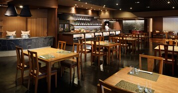 A restaurant with wooden tables and chairs, a bar area with bottles, and a chef preparing food.