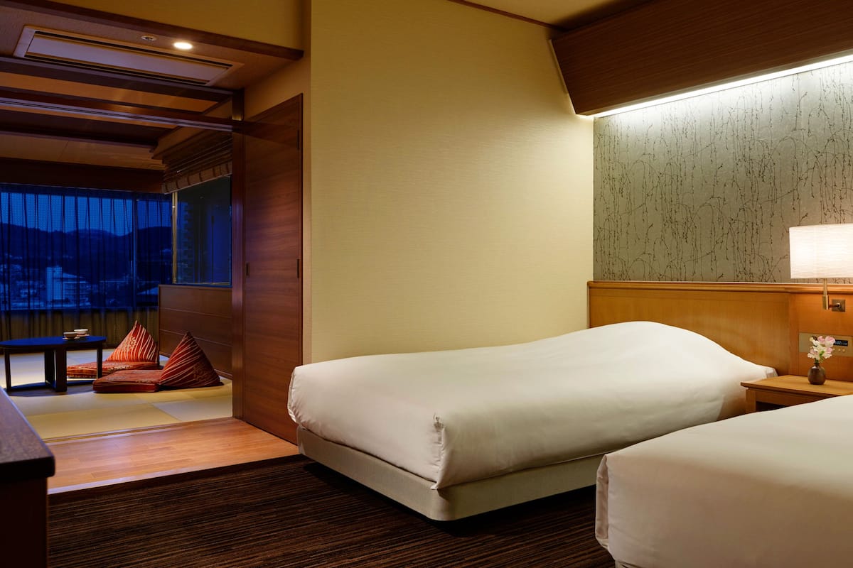 A hotel room with two beds, a wooden headboard, a nightstand, and a window with a view of mountains.