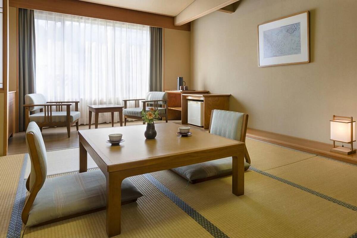 Une pièce de style japonais traditionnel, avec un sol recouvert de tatamis, une petite table sur laquelle sont posés un bol et un vase, et un bureau avec une lampe.