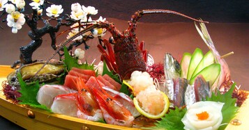 Variedad de mariscos dispuestos en un plato de madera, incluyendo camarones, pescado y otras exquisiteces, con un árbol decorativo y flores al fondo.