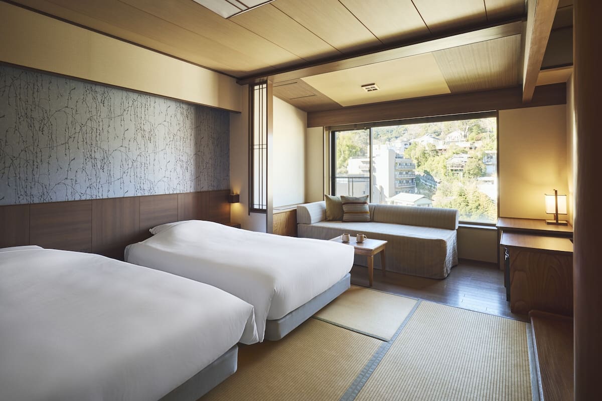 A hotel room with two beds, a sofa, a small table, and a window with a view of buildings.
