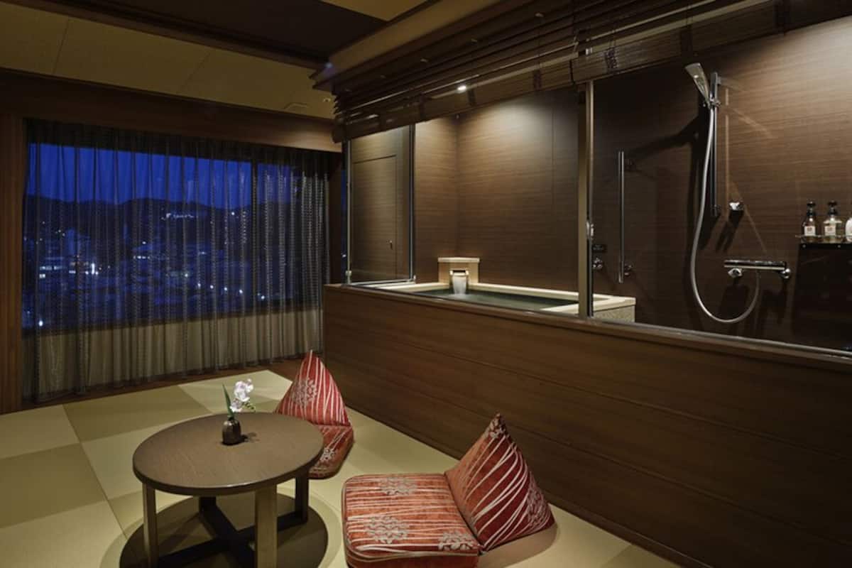 A hotel room with a large window, a sofa, a small table, and a shower area.