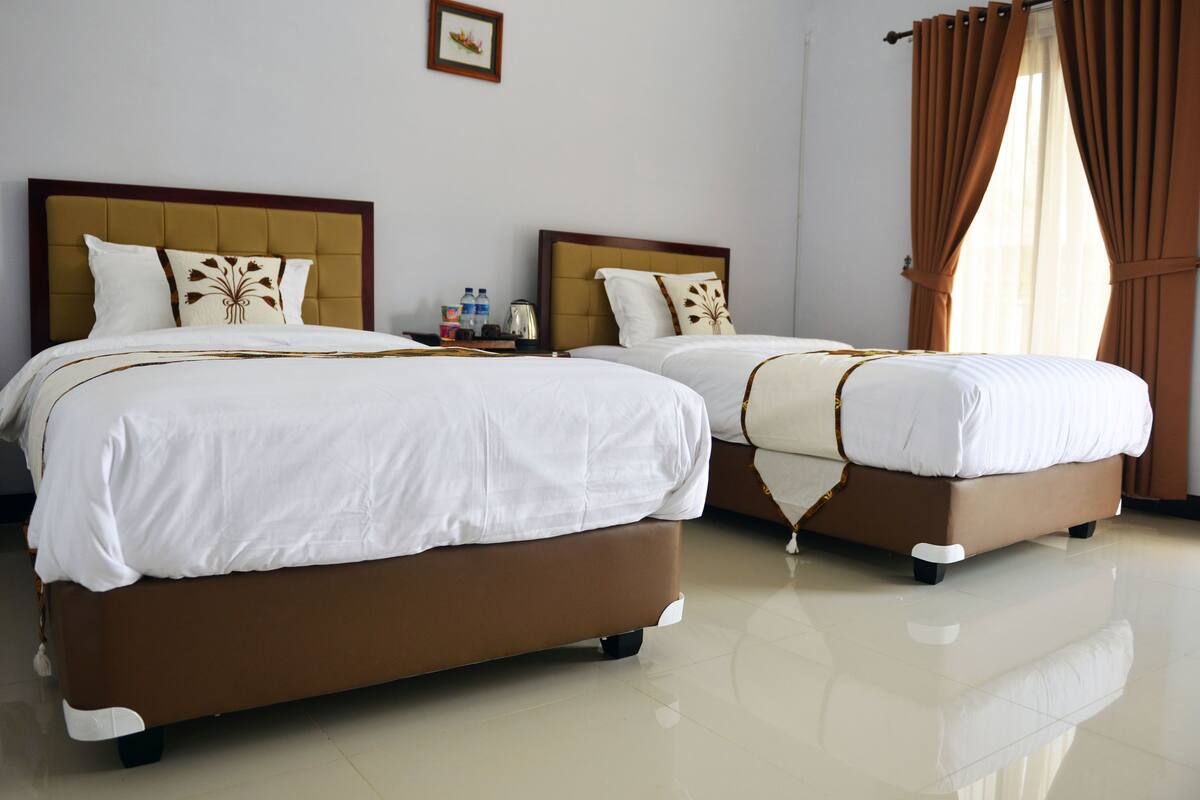 A hotel room with two beds, each with a headboard and pillows. There is a small table with a water bottle and a cup.