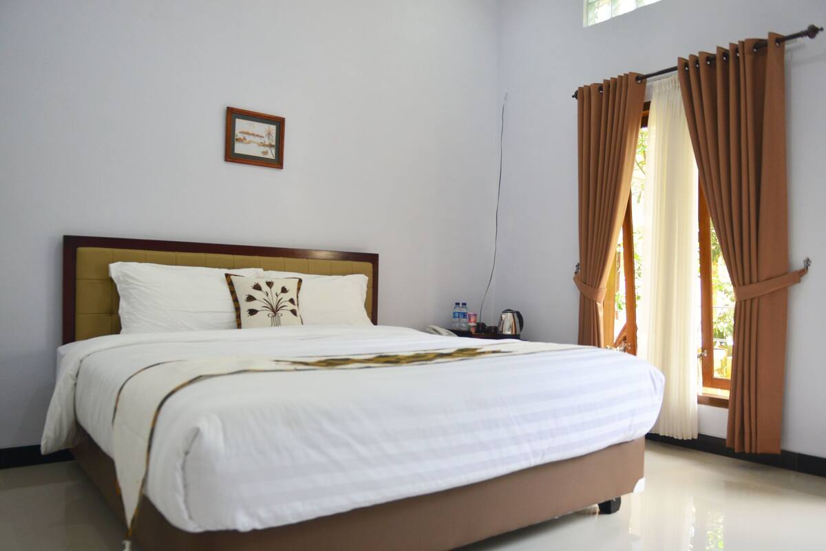 A neatly made bed with white linens and a decorative pillow, a bedside table with a water bottle and a kettle, and a window with brown curtains.