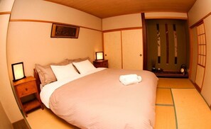 A traditional Japanese-style bedroom with tatami flooring, a low bed with a white blanket, a wooden nightstand with a lamp, and a sliding door with decorative paper. - Morino Lodge Hakuba (Hakuba)