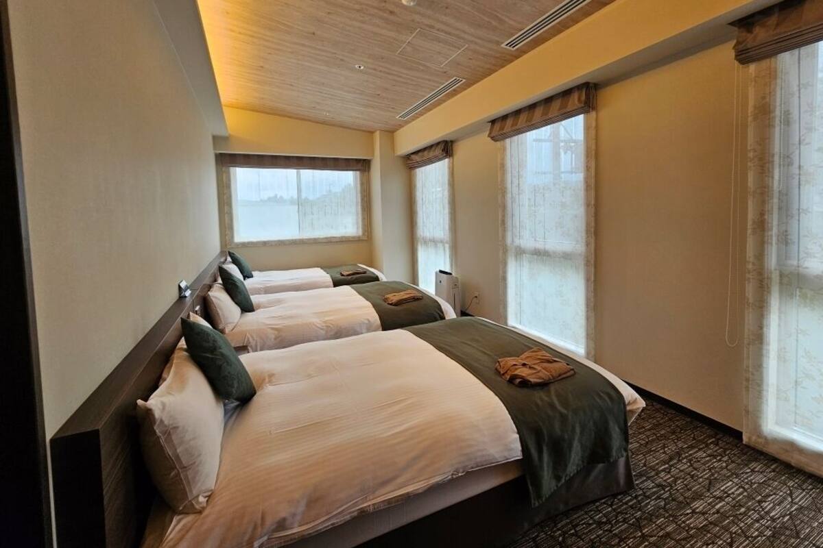 Une chambre d’hôtel avec trois lits, de grandes fenêtres et un plafond en bois.