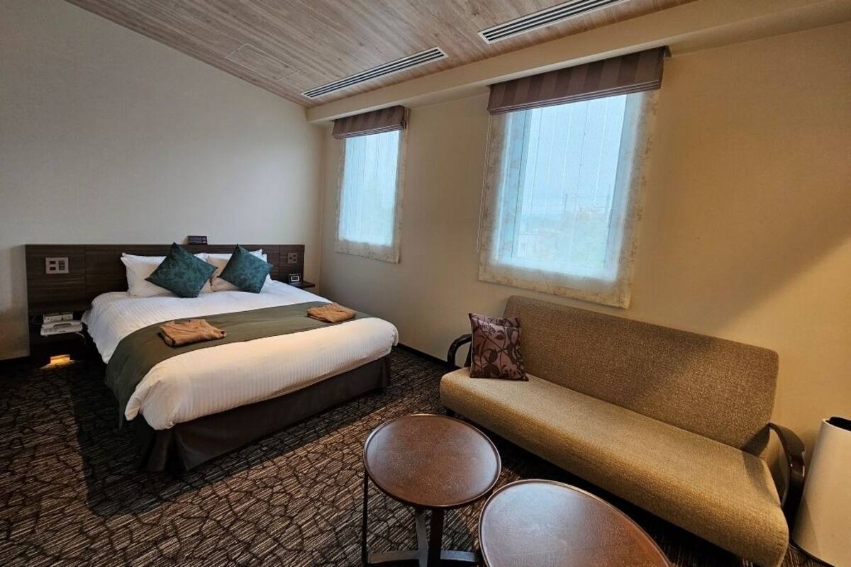Une chambre d’hôtel avec un lit, un canapé, deux petites tables et deux fenêtres.