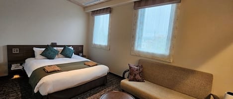 A hotel room with a bed, a sofa, two small tables, and two windows.