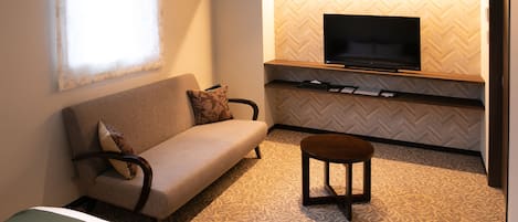 Ein gemütlicher Wohnraum mit Sofa, kleinem Tisch, Flachbildfernseher an einer Wand mit Fischgrätmuster und Fenster mit transparenten Vorhängen.