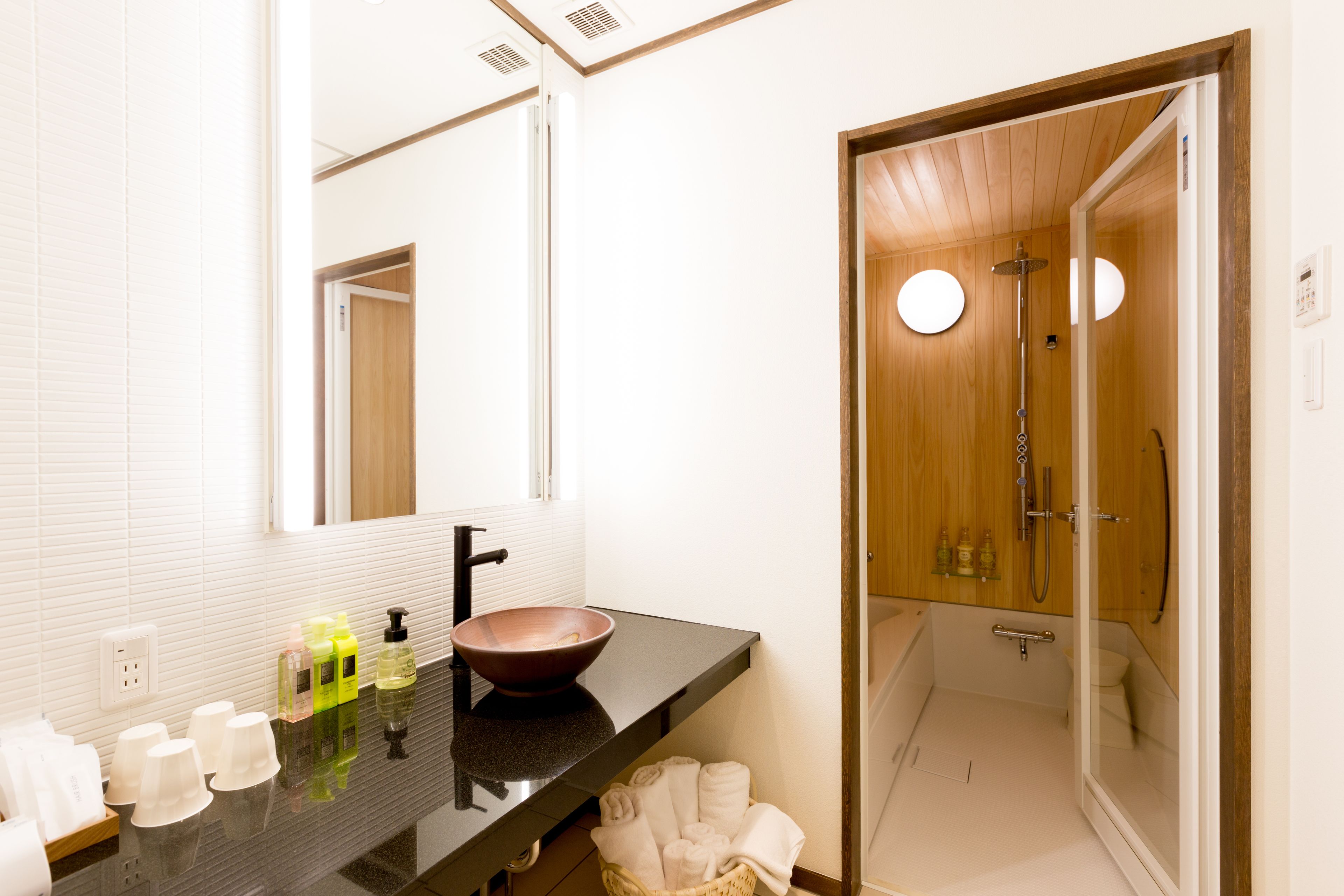 A modern bathroom with a black countertop, a large mirror, a wooden door leading to a shower area, and a basket of towels.