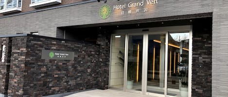 Exterior del Hotel Grand Vert, con fachada de ladrillo moderno, grandes puertas de entrada de vidrio y un cartel con el nombre del hotel.