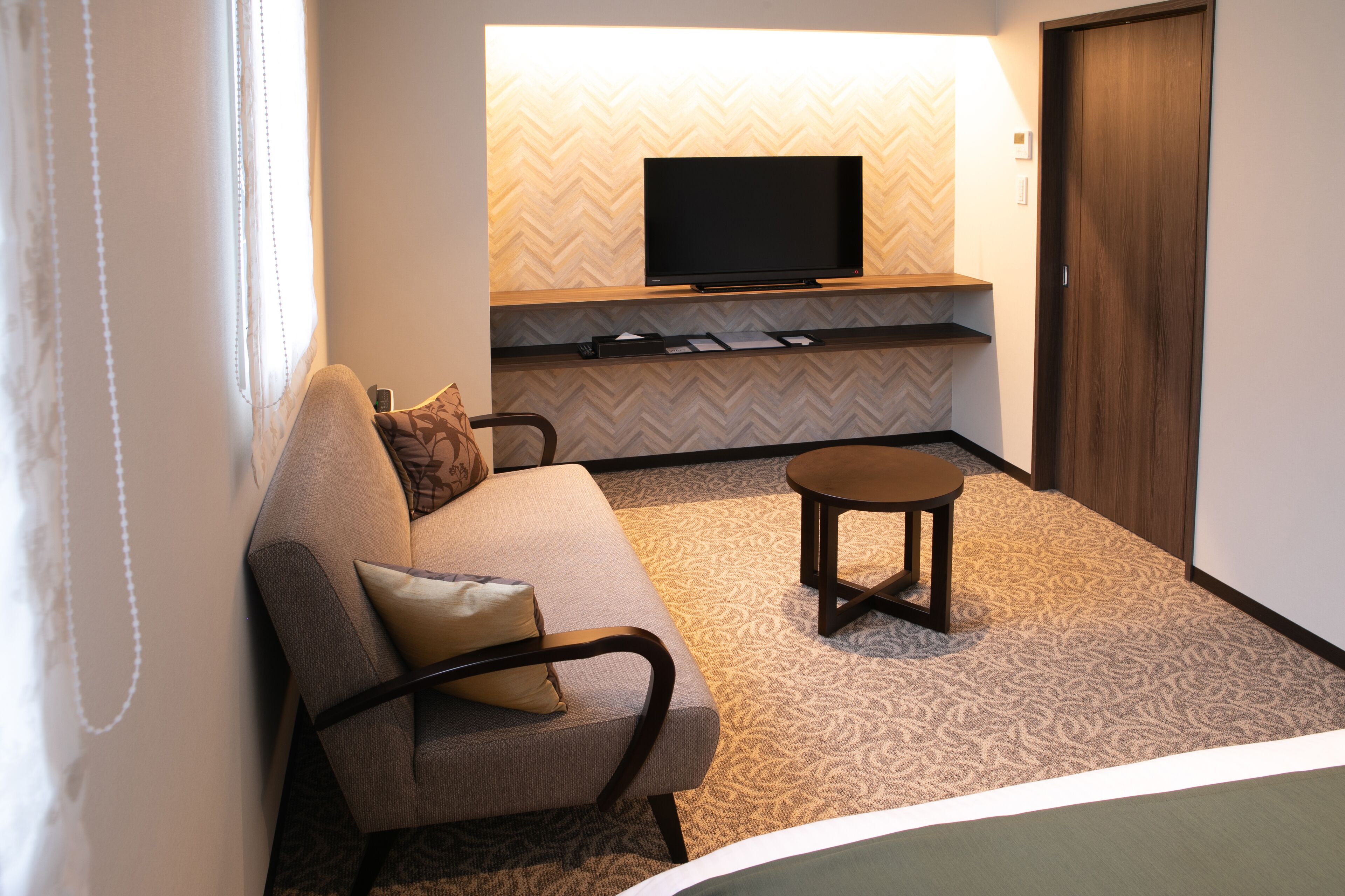 A hotel room with a sofa, a small table, a television on a wooden shelf, and a patterned carpet.