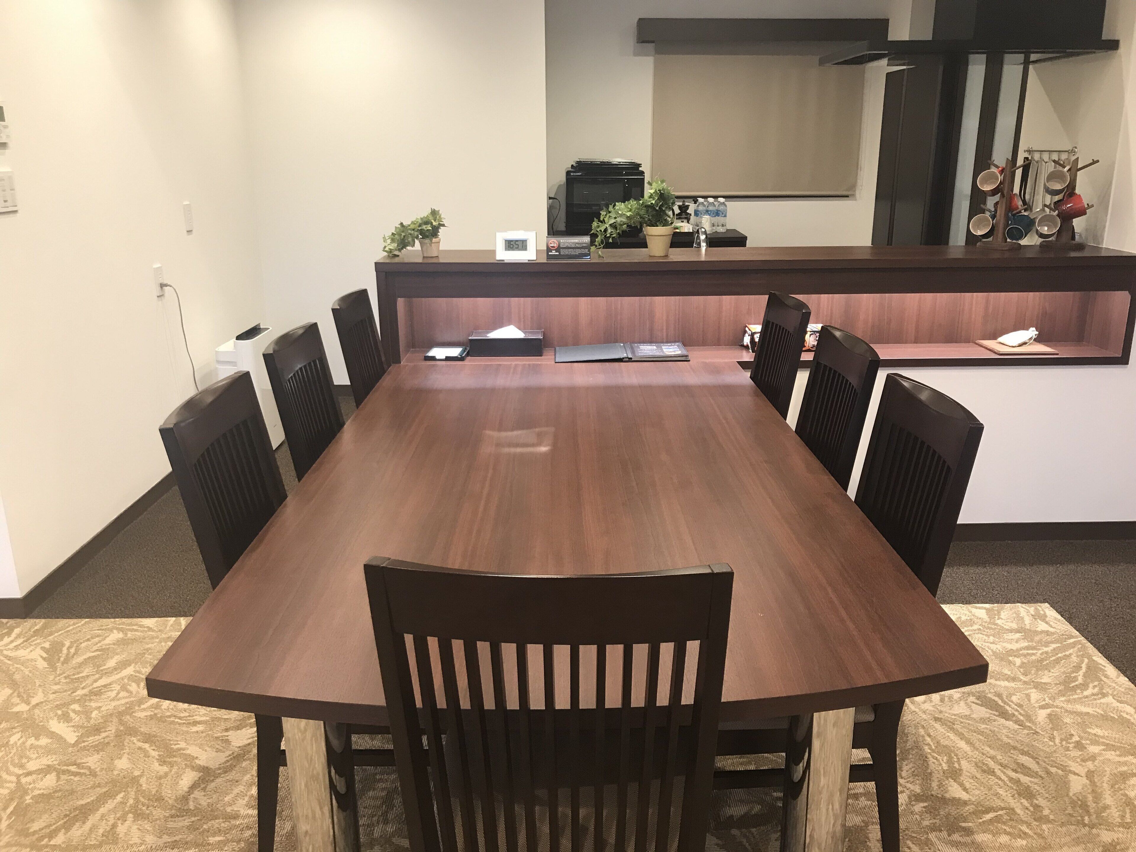 Una sala de conferencias con una larga mesa de madera, ocho sillas, una pequeña zona de cocina con un microondas y algunas plantas en macetas.
