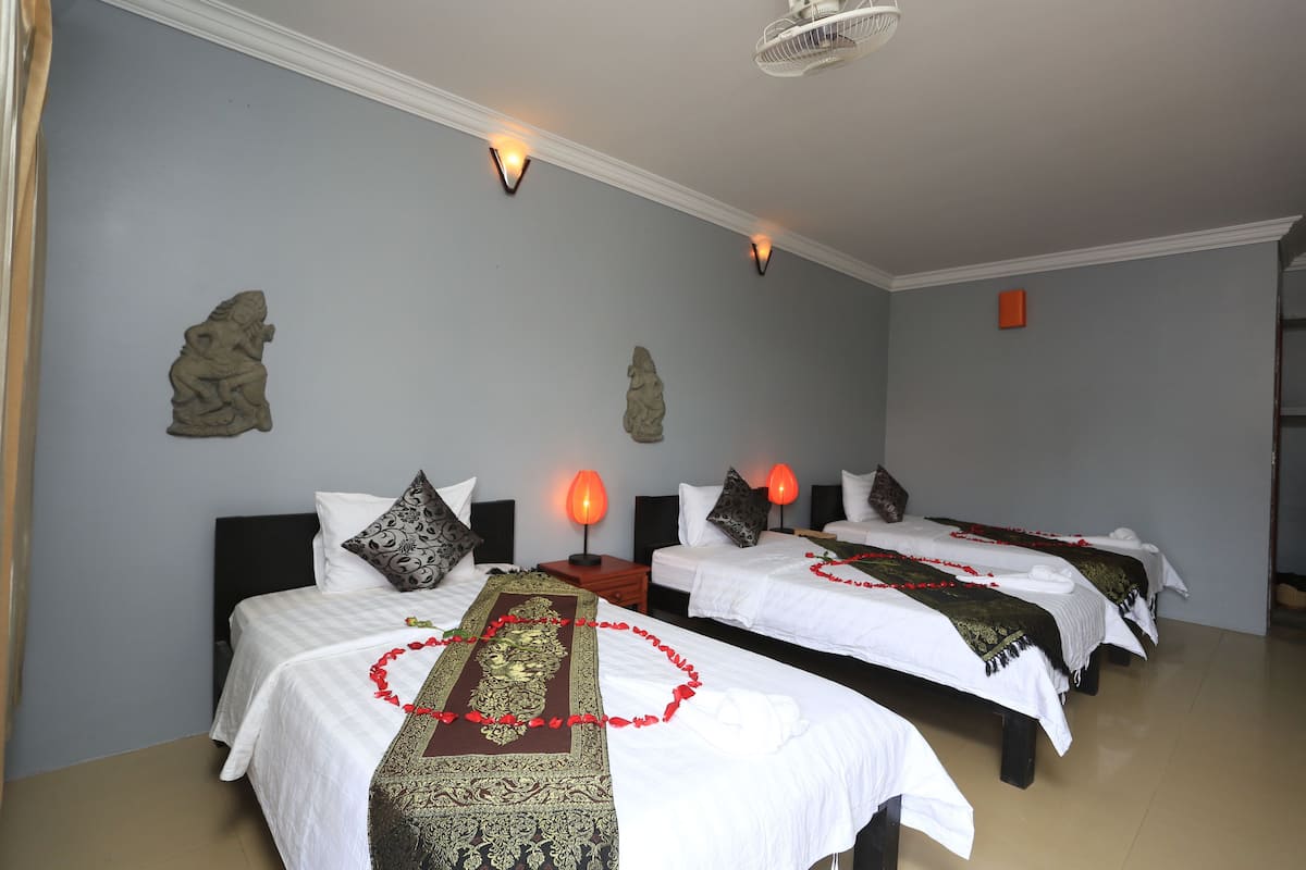 Quarto de hotel com duas camas, cada uma com roupa de cama branca e almofadas decorativas. Há arandelas na parede que fornecem luz, e um ventilador de teto é visível.