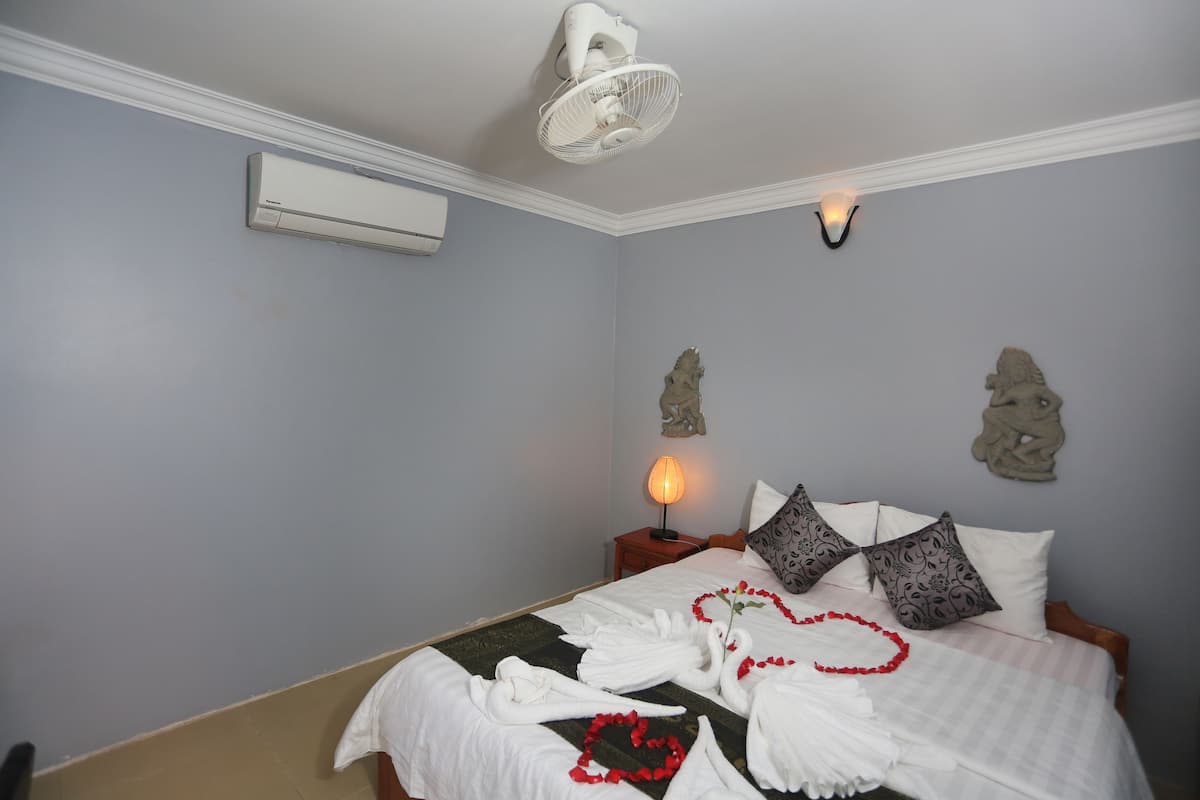 Uma cama bem arrumada com lençóis brancos, um objeto decorativo na parede, um ar condicionado fixado na parede e um ventilador de teto.