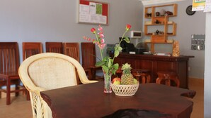 A dining area with a wooden table, wicker chairs, a fruit basket, and a bulletin board in the background. - Cheng Lay Guesthouse (Siem Reap)