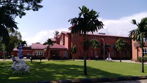 A red building with multiple windows and a red roof, surrounded by green grass and palm trees. - Myanmar Sports Hotel (Yangon)